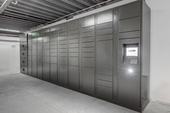 A row of tall, dark grey lockers are lined up in a room with a concrete floor and a white wall.